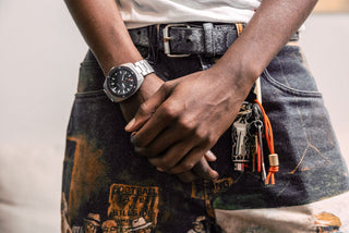 Person wearing a watch with a belt and keys, standing against a neutral background.