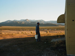 Artist Jess Mudgett holding a bucket of paint while staring at a blank wall with the desert mountains in the background.