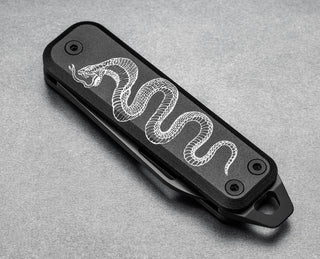 The Schoph collab Elko pocket knife with a snake graphic closed on a steel surface.