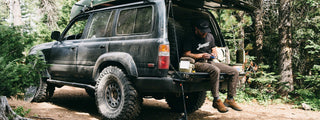 A man on the tailgate of his SUV in the forest.