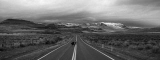 Two guys skateboarding down a lonely highway towards snow capped hills.