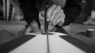A man cutting out a leash plug on a surfboard with a pocket knife.