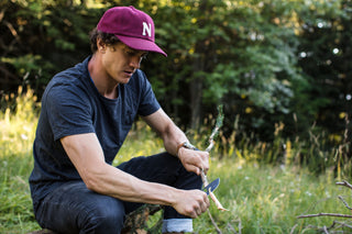 A man shaving a stick for a fire with his pocekt knife.