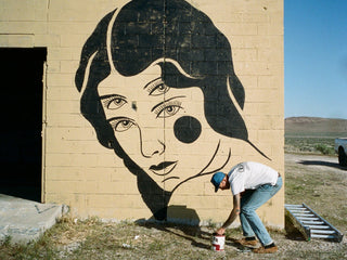 Artist Jess Mudgett painting a four eyed woman on a wall in the desert.