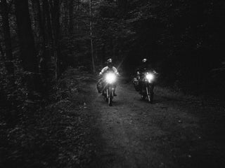 Two motorcyclist riding in the dark down a dirt road in the wilderness.